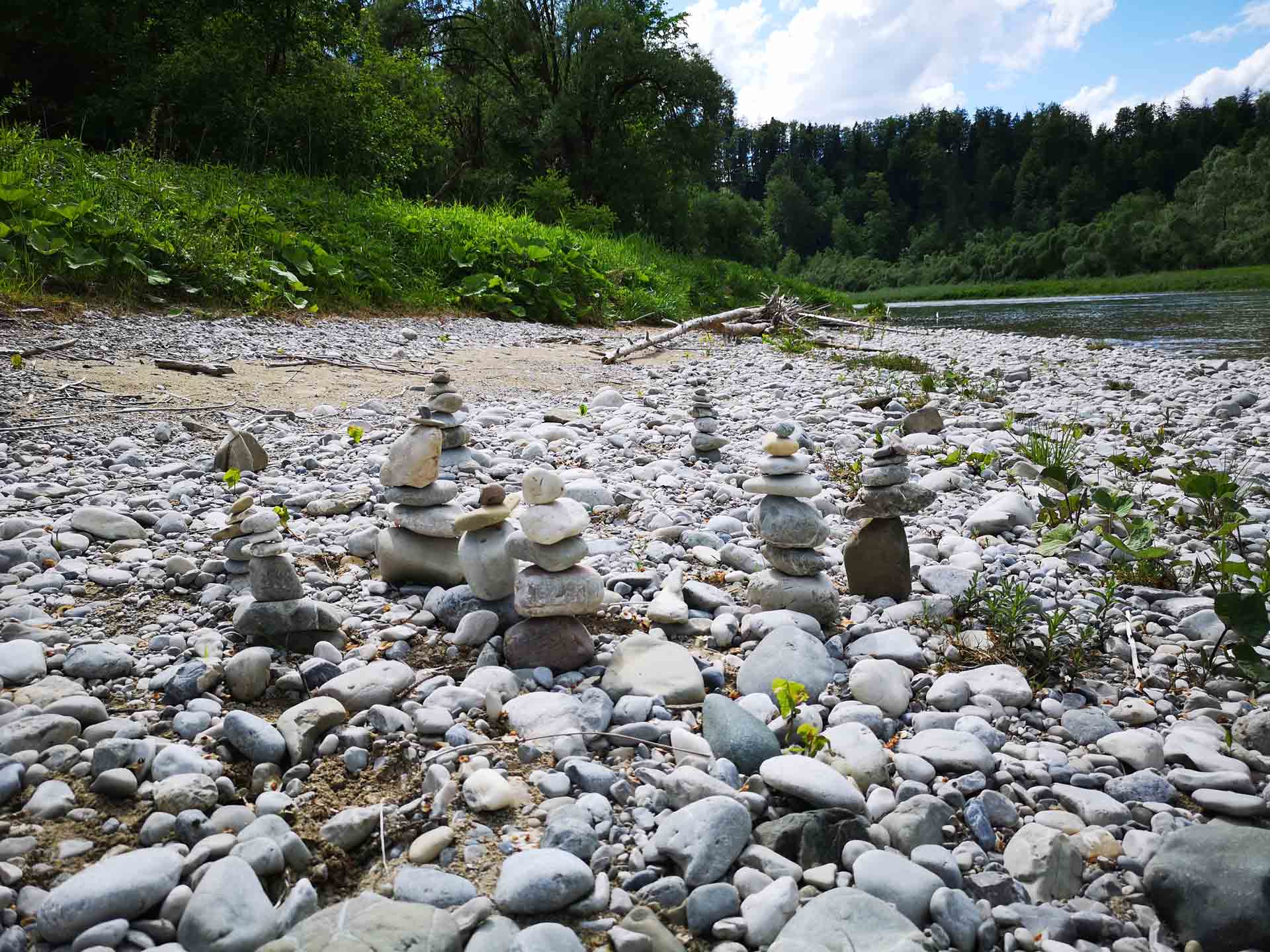 Mehrere sorgfältig gestapelte Steintürme auf einer breiten Schotterbank am Flussufer.