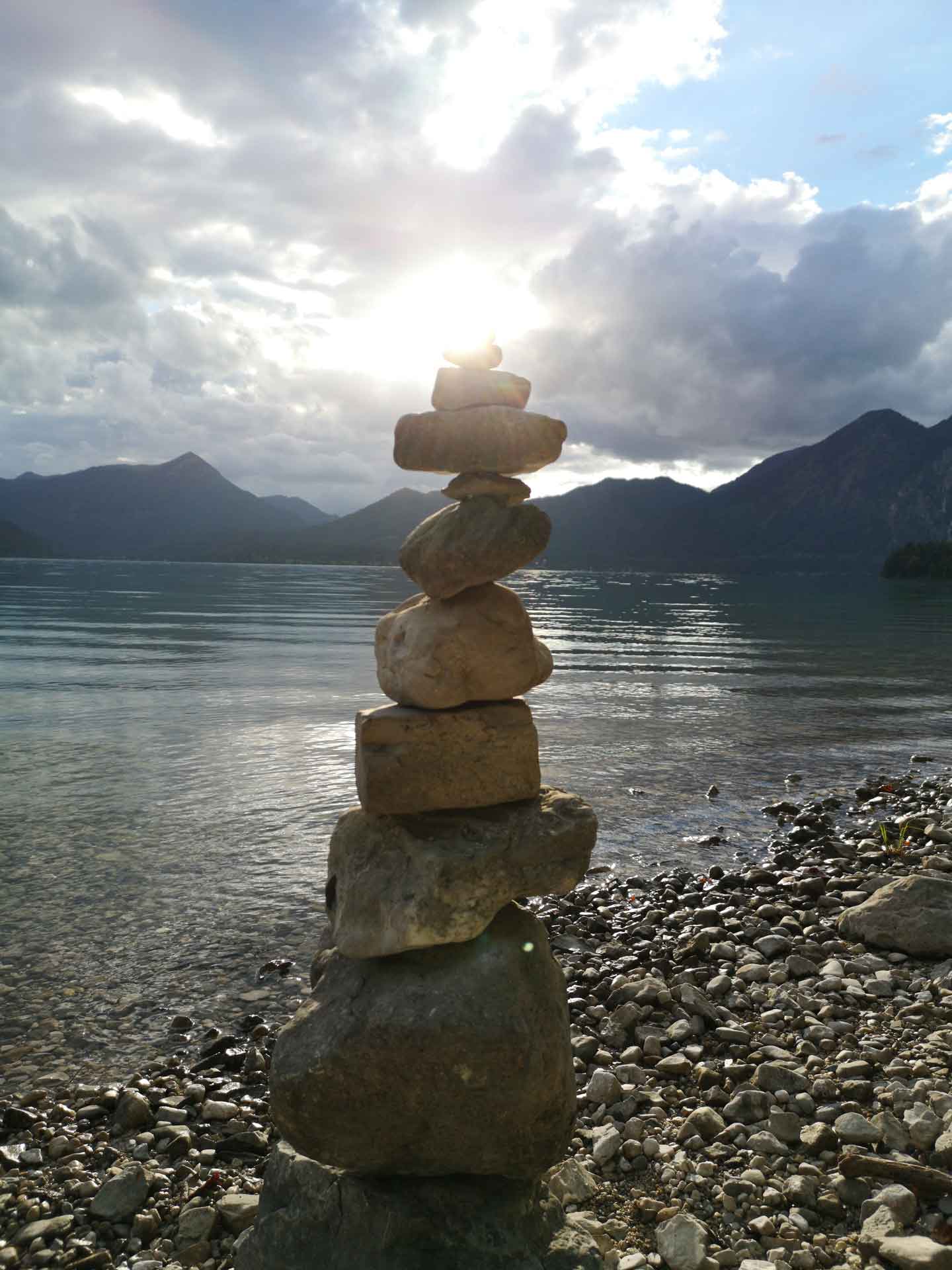 Steinmandl am steinigen Ufer eines Bergsees; die Sonne scheint durch die Wolken.
