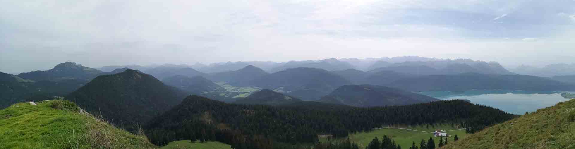 Weitwinkel-Panorama der Bayerischen Alpenkette mit grünen Wäldern, Almwiesen und türkisfarbenem Bergsee.
